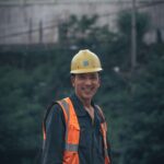 A smiling construction worker wearing safety gear stands outdoors in a work environment.